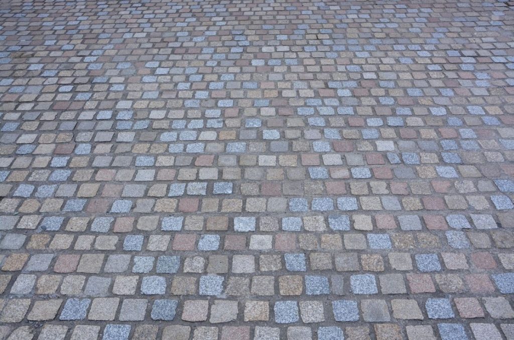 Deze afbeelding toont een oprit of bestrating die is aangelegd met natuurstenen of gebakken klinkers in een blokpatroon. De stenen hebben verschillende tinten – grijs, beige, roodachtig – wat wijst op het gebruik van oude of gebruikte klinkers, die bekend staan om hun duurzaamheid en karakter.