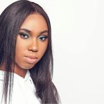 Deze afbeelding toont een vrouw met lang, stijl haar en opvallende oogmake-up in groene tinten. Ze draagt een witte hoodie en staat tegen een witte achtergrond, wat zorgt voor een minimalistische, professionele uitstraling.