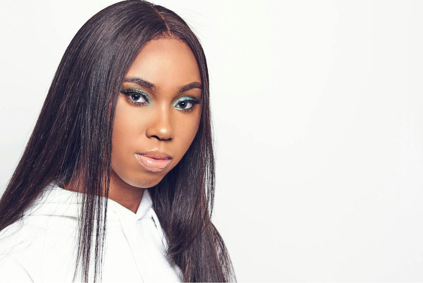 Deze afbeelding toont een vrouw met lang, stijl haar en opvallende oogmake-up in groene tinten. Ze draagt een witte hoodie en staat tegen een witte achtergrond, wat zorgt voor een minimalistische, professionele uitstraling.