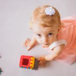 Peuter in een zachtroze jurk speelt op de vloer met kleurrijke blokken in een ruimte met warme kleurpaletten.