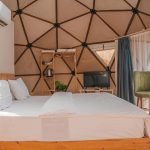 Glamping slaapkamer met houten interieur, airco en geometrisch koepeldak, voorzien van wit bed en TV.