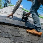 Een dakdekker plaatst asfaltshingles met een spijkerpistool op een schuin dak onder een heldere lucht.