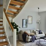 Moderne woonkamer met open houten trap en lichtgrijze bank onder een witte muur met kunst