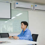 Man in vergaderruimte onder drie wandgemonteerde airco-units, zittend aan tafel met afstandsbediening